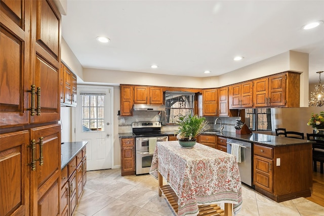 kitchen with brown cabinetry, dark stone counters, stainless steel appliances, under cabinet range hood, and backsplash