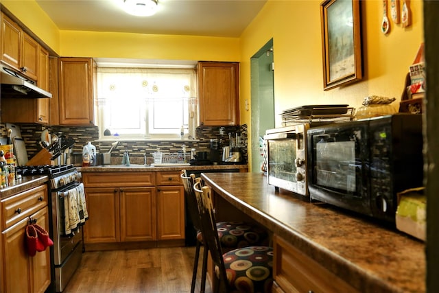 kitchen featuring decorative backsplash, brown cabinetry, dark countertops, under cabinet range hood, and stainless steel range with gas stovetop