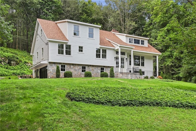 view of front of home featuring a shingled roof, a front yard, stone siding, and a chimney