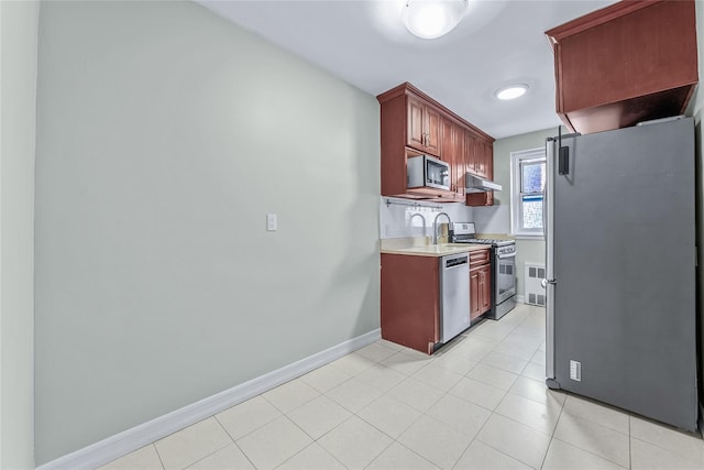 kitchen with under cabinet range hood, stainless steel appliances, baseboards, light countertops, and reddish brown cabinets