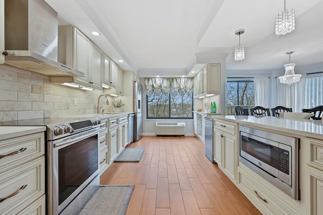 kitchen with stainless steel appliances, a sink, hanging light fixtures, wall chimney range hood, and cream cabinetry