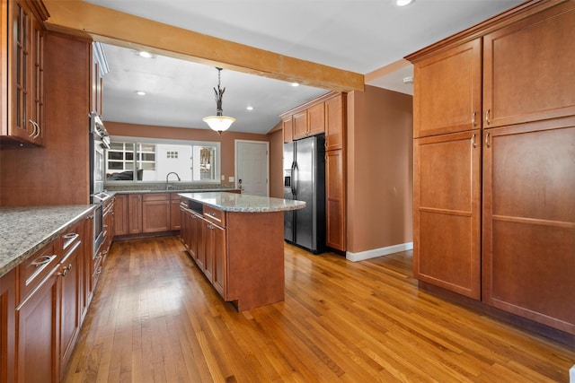 kitchen with light stone counters, light wood finished floors, brown cabinetry, a kitchen island, and stainless steel fridge