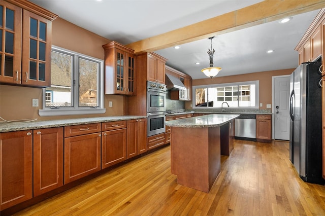 kitchen with light stone counters, stainless steel appliances, glass insert cabinets, a kitchen island, and wall chimney exhaust hood