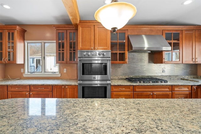 kitchen featuring light stone countertops, stainless steel appliances, decorative backsplash, wall chimney exhaust hood, and glass insert cabinets