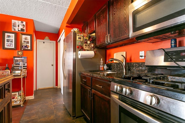 kitchen with appliances with stainless steel finishes, a sink, a textured ceiling, dark stone counters, and baseboards