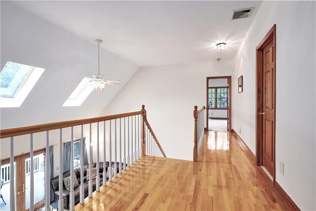 hallway with lofted ceiling with skylight, visible vents, light wood-style flooring, and an upstairs landing