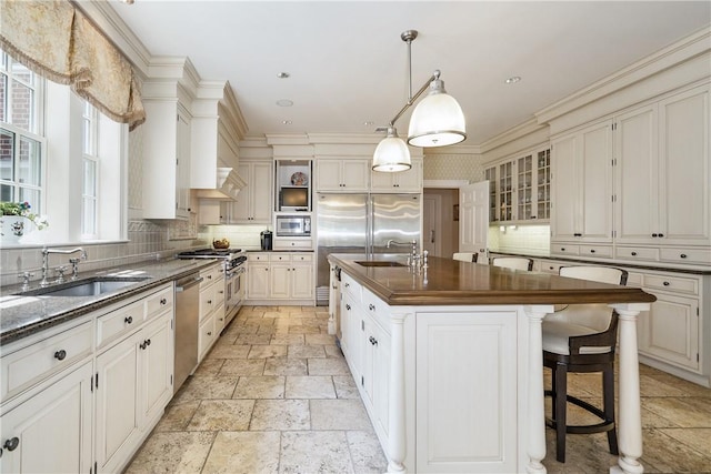 kitchen with a kitchen island with sink, stainless steel appliances, a sink, glass insert cabinets, and pendant lighting