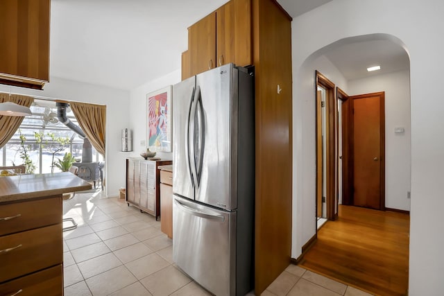 kitchen featuring arched walkways, light tile patterned floors, light countertops, freestanding refrigerator, and brown cabinets