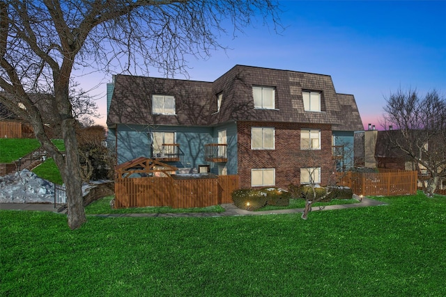 rear view of property featuring brick siding, a lawn, fence, and mansard roof