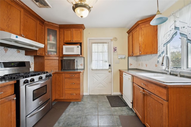 kitchen featuring under cabinet range hood, white appliances, a sink, brown cabinetry, and glass insert cabinets