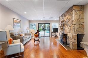 living area with a drop ceiling, a stone fireplace, and wood finished floors