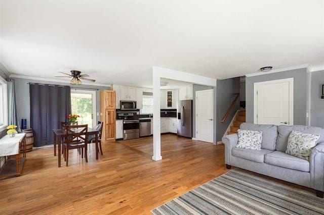 living room featuring light wood finished floors, stairs, a ceiling fan, and crown molding