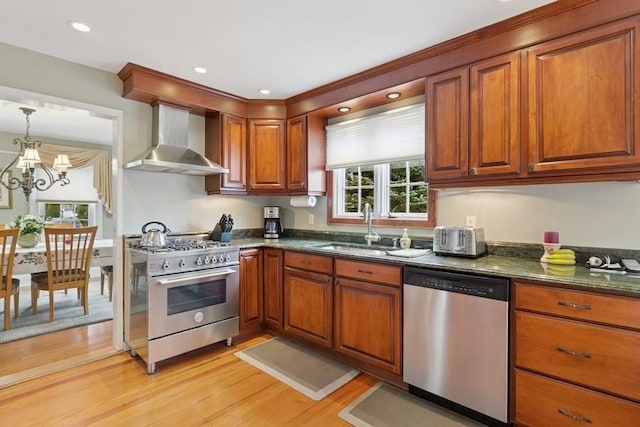 kitchen featuring brown cabinetry, dark stone counters, appliances with stainless steel finishes, wall chimney range hood, and a sink