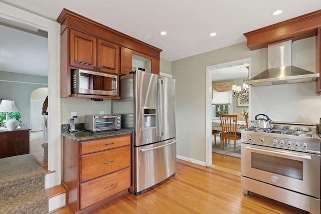 kitchen featuring arched walkways, stainless steel appliances, light wood-style floors, wall chimney range hood, and brown cabinetry