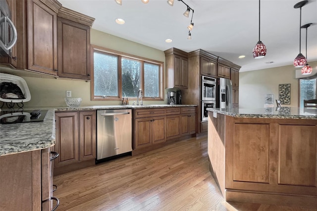 kitchen featuring brown cabinetry, light stone counters, hanging light fixtures, stainless steel appliances, and light wood-type flooring