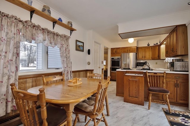 kitchen featuring a sink, stainless steel appliances, a peninsula, wainscoting, and brown cabinetry