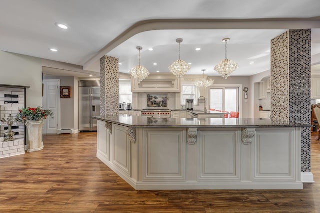 kitchen featuring dark wood-style flooring, built in refrigerator, decorative backsplash, dark stone counters, and decorative light fixtures