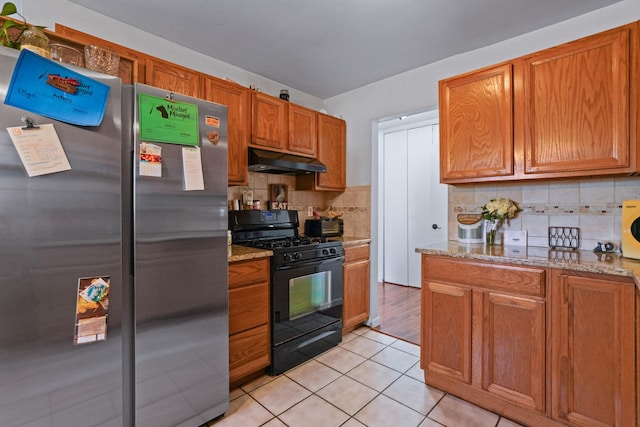 kitchen with under cabinet range hood, backsplash, freestanding refrigerator, light stone countertops, and gas stove