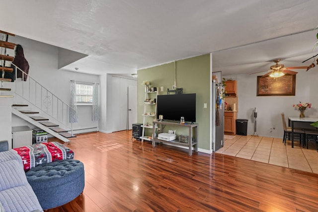 living area with baseboard heating, a ceiling fan, wood finished floors, baseboards, and stairs