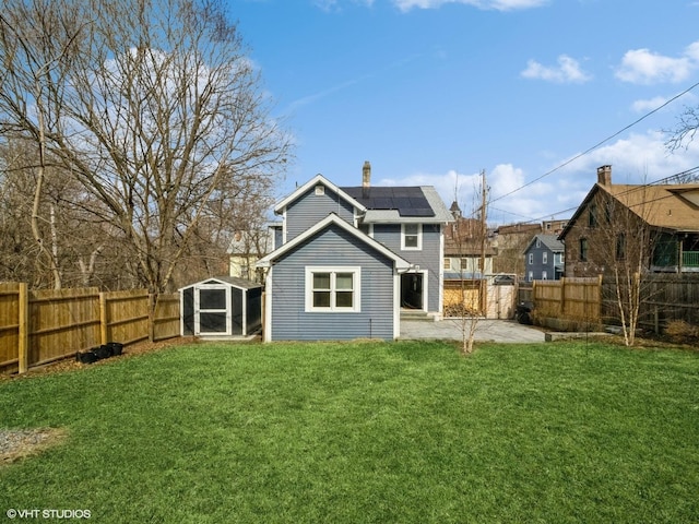 back of house featuring a patio, a fenced backyard, a lawn, roof mounted solar panels, and a chimney