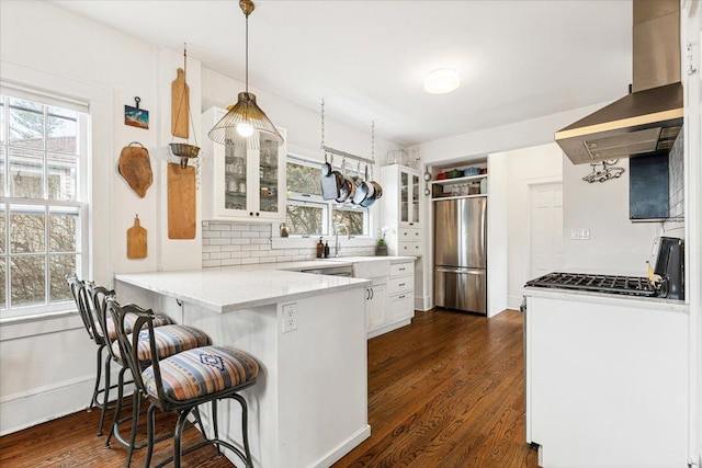 kitchen featuring stainless steel refrigerator, white cabinetry, a peninsula, wall chimney range hood, and glass insert cabinets