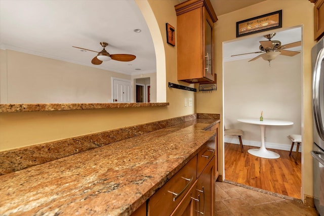 kitchen featuring baseboards, brown cabinetry, ceiling fan, ornamental molding, and stone counters