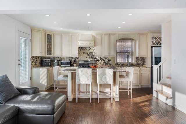 kitchen featuring cream cabinets, stainless steel appliances, a breakfast bar, glass insert cabinets, and custom range hood