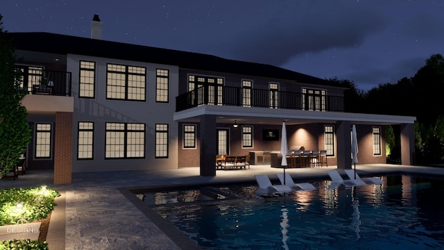 back of house at night featuring a balcony, brick siding, an outdoor pool, and a patio