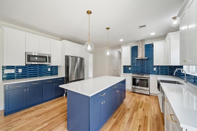 kitchen with blue cabinets, a kitchen island, light countertops, appliances with stainless steel finishes, and wall chimney range hood