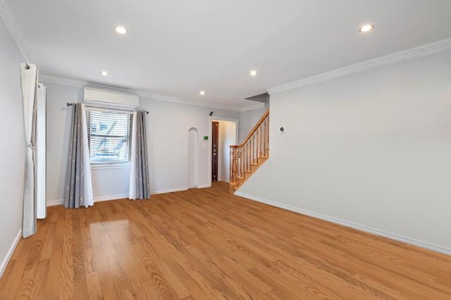 empty room featuring crown molding, light wood-style flooring, a wall mounted air conditioner, baseboards, and stairs