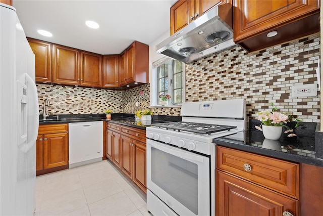 kitchen with tasteful backsplash, brown cabinetry, dark stone countertops, white appliances, and under cabinet range hood