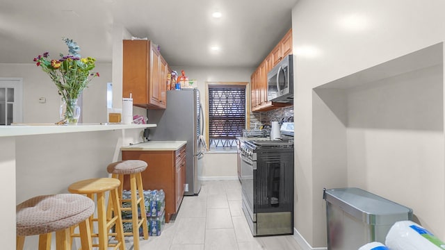 kitchen featuring a breakfast bar area, stainless steel appliances, light countertops, decorative backsplash, and brown cabinetry