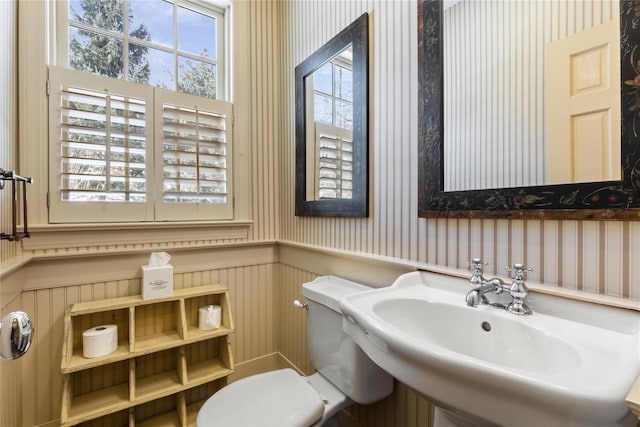 bathroom with toilet, wallpapered walls, a sink, and wainscoting