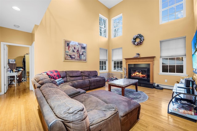 living room featuring a high ceiling, baseboards, light wood-style floors, baseboard heating, and a glass covered fireplace