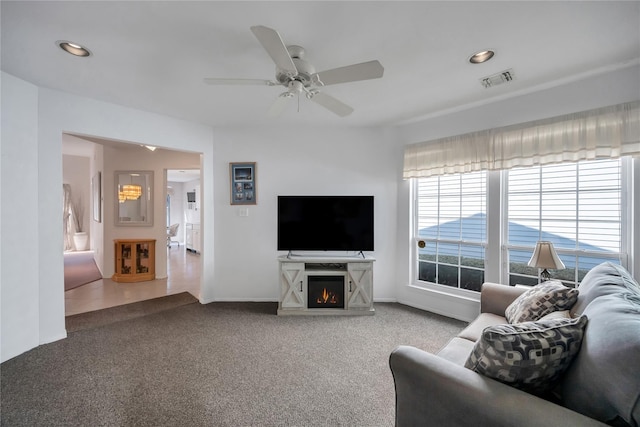 carpeted living area with recessed lighting, a warm lit fireplace, visible vents, and ceiling fan