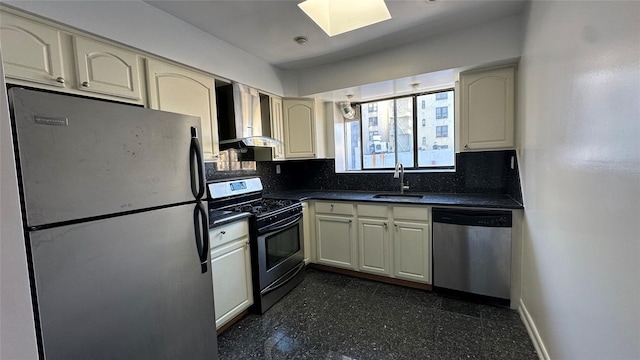 kitchen with stainless steel appliances, dark countertops, cream cabinets, a sink, and wall chimney exhaust hood