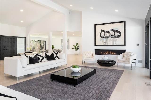 living area with light wood finished floors, recessed lighting, visible vents, high vaulted ceiling, and a warm lit fireplace