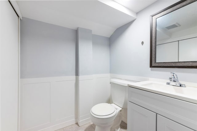 half bathroom with toilet, a decorative wall, vanity, visible vents, and wainscoting