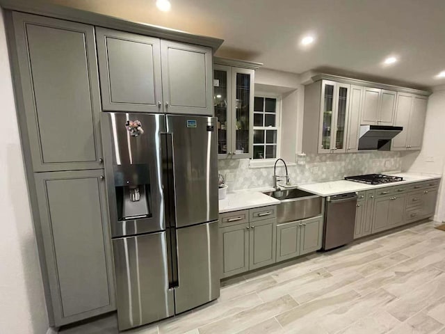 kitchen with stainless steel appliances, backsplash, glass insert cabinets, a sink, and under cabinet range hood