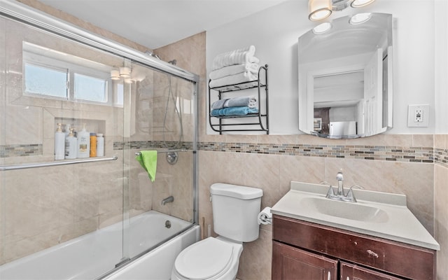 bathroom featuring wainscoting, toilet, shower / bath combination with glass door, vanity, and tile walls