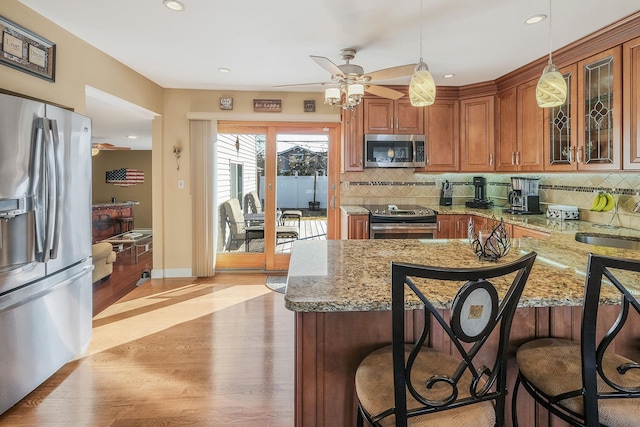 kitchen featuring brown cabinets, decorative light fixtures, stainless steel appliances, glass insert cabinets, and light stone countertops