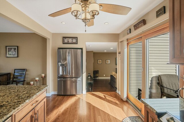 kitchen with stainless steel fridge with ice dispenser, ceiling fan, brown cabinets, wood finished floors, and stone counters