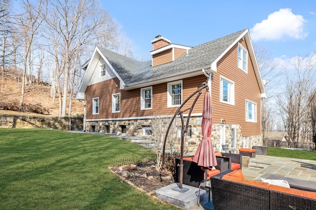 back of property with a patio, stone siding, a lawn, roof with shingles, and a chimney