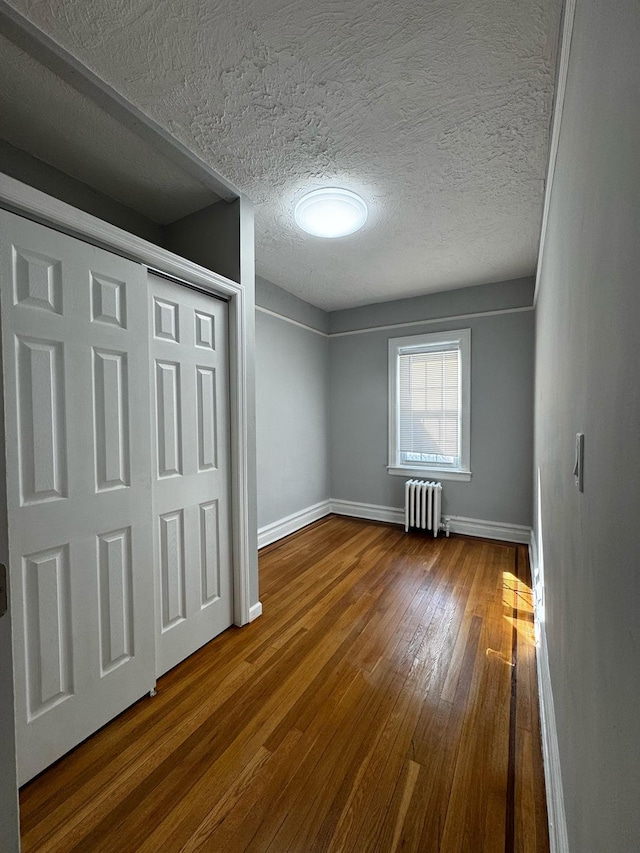 unfurnished bedroom with a textured ceiling, wood finished floors, baseboards, a closet, and radiator