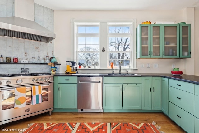 kitchen featuring dark countertops, appliances with stainless steel finishes, glass insert cabinets, a sink, and wall chimney exhaust hood