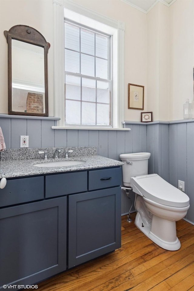 half bath with toilet, wood finished floors, vanity, ornamental molding, and wainscoting