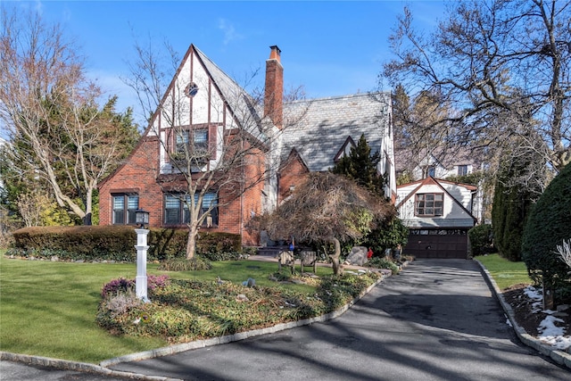 tudor home featuring an attached garage, a chimney, a front lawn, and brick siding