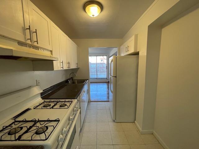 kitchen with light tile patterned floors, under cabinet range hood, white appliances, a sink, and white cabinetry