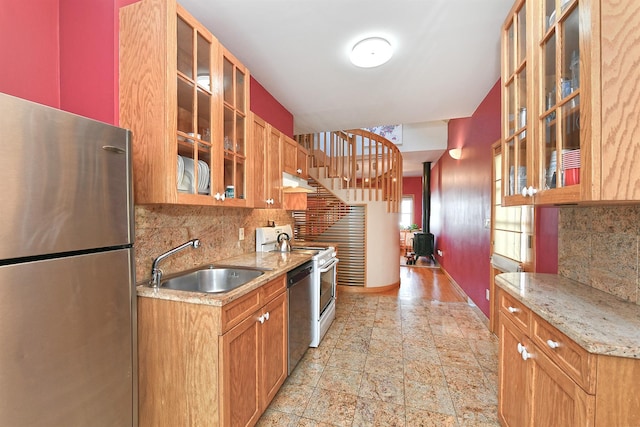 kitchen featuring stainless steel appliances, brown cabinetry, glass insert cabinets, a sink, and light stone countertops