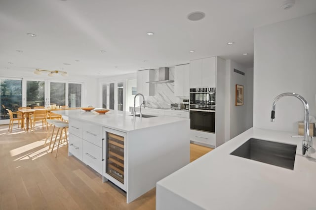 kitchen with beverage cooler, a sink, white cabinetry, wall chimney range hood, and a center island with sink